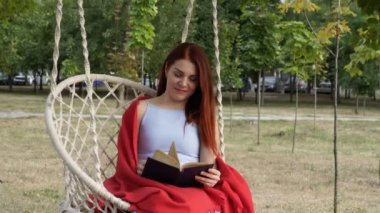 Kızıl saçlı genç bir kız, şehir parkında beyaz asılı salıncakta oturan bir kitap okuyor. Güneşli bir sonbahar gününde kırmızı bir battaniyeyle kaplı olarak gülümsüyor ve hayal ediyor. Portre. Close. 4k.