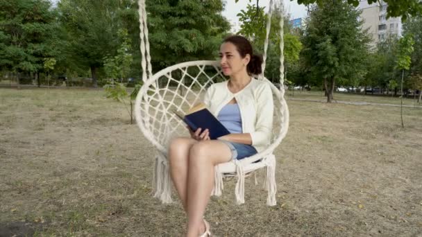 Een oosterse vrouw van middelbare leeftijd ligt in een stadspark en schommels op een witte schommel. Ze leest een boek en ziet er dromerige rond op een zonnige herfst dag. Portret. Close-up. 4k. — Stockvideo