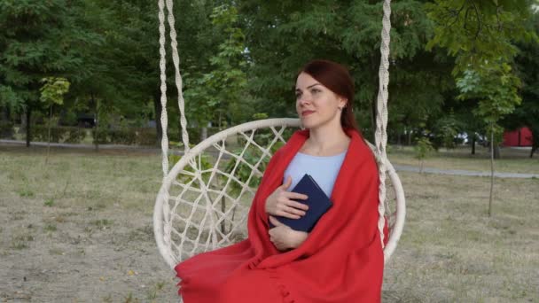 Una bella ragazza dai capelli rossi si siede con un libro in mano su un'altalena nel parco al tramonto di una giornata estiva, coperta da un plaid rosso. Sospira sognante e sorride. Primo piano. 4K . — Video Stock