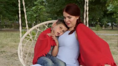 Gün batımında Park'ta salıncakta mutlu bir anne ve bebeğin portresi. Kızıl saçlı bir kız, küçük oğlunu kucağına tutar, ona sarılır ve kırmızı bir battaniye yle onu örter. Yakından kapatın. 4k.