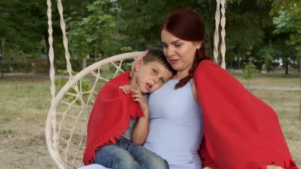 Portrait d'une maman et d'un bébé heureux sur une balançoire dans le parc au coucher du soleil. Une fille aux cheveux roux tient son jeune fils sur ses genoux, le serre dans ses bras et le couvre d'une couverture rouge. Ferme là. 4K . — Video