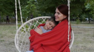 Mutlu bir anne kucağında küçük oğlu tutar ve gün batımında parkta bir salıncak üzerinde otururken onu beşik. O, onu okşayarak ve okşayarak, kırmızı battaniye altında onu kucaklar. Kavram. Close. 4k.