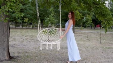 Güzel kız modeli moda fotoğrafçılığı için açık havada poz. O duruyor ve gün batımında şehir Park'ta beyaz salıncak seti yakınında mavi bir elbise saçlarını düzeltir. Moda portresi. 4k.