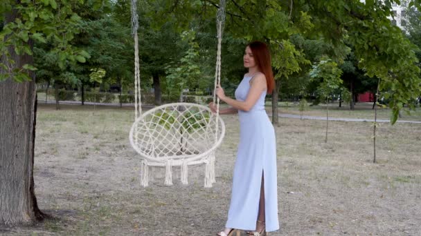 Modelo profissional, uma menina com cabelo vermelho, posando para fotografia de moda ao ar livre. Ela fica em um vestido azul perto do balanço branco no parque da cidade ao pôr-do-sol. Retrato de moda. 4K . — Vídeo de Stock