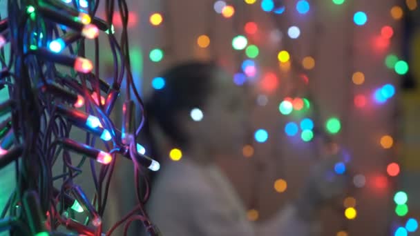 Close-up colorful blinking Christmas garland on a blurred background. Beautiful bokeh. Light spot. A blurry child plays in the background. Festive Christmas illuminations. Shallow depth of field. 4K. — Stock Video