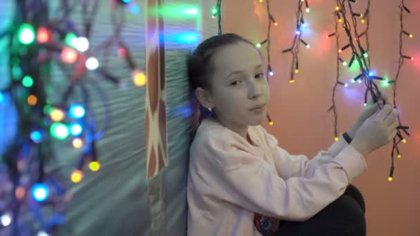 Portrait of a sad little girl on the background of flashing colorful Christmas lights. She sits among the small, colorful garlands, and looking at the camera. Shallow depth of field. Close-up. 4K. — Stock Video