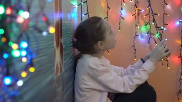 Niña pensativa sentada contra la pared y jugando con coloridas guirnaldas de Navidad. Toca las pequeñas luces intermitentes y sonríe tristemente. Iluminación festiva. Pequeña profundidad de campo. 4K . — Vídeo de stock