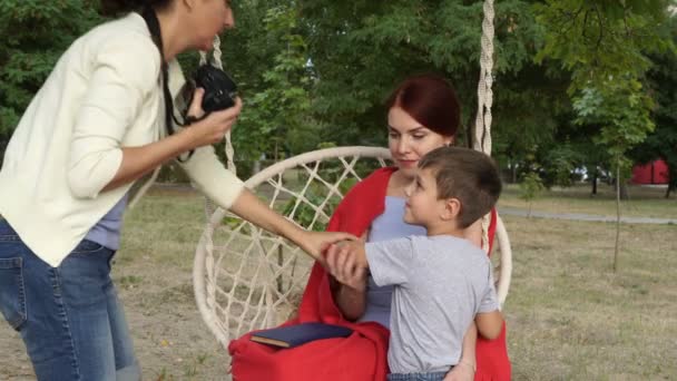 Східна дівчина фотограф стріляє мамі і дитині в парк на гойдалці. Вони ставлять для неї сидить на білому гойдалці, покриті червоною ковдру на заході сонця. Фотограф на роботі. Закри. 4K. — стокове відео