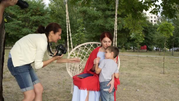 Два профессиональных фотографа разных полов снимают красивую семью на свежем воздухе, на качелях в городском парке. Они объясняют и показывают матери и ребенку, как позировать. Семейная фотография. 4K . — стоковое видео