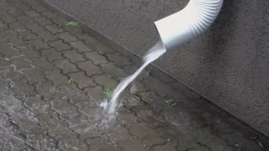 Evin üzerindeki beyaz bir drenaj borusu, kentte yağmur sırasında kaldırım lara bir su akışı döküyor. Üst yan görünüm. 4k