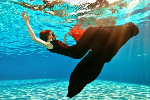 Una chica inusual se ahoga en la piscina. Una chica con el pelo rojo y un vestido cae al fondo de la piscina en el fondo de la luz del sol. Surrealismo. Concepto . — Foto de Stock