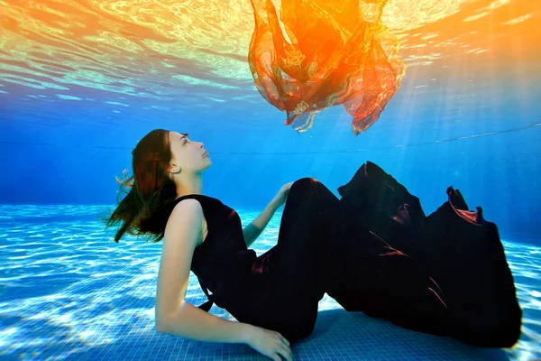 Een jong meisje verdrinkt en kijkt omhoog vanaf de bodem van het zwembad. Ze ligt aan de onderkant in een modieuze Bourgondische jurk op een achtergrond van gele vulling van het oppervlak. Surrealisme. Portret. Concept — Stockfoto