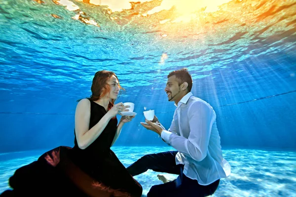 Ein ungewöhnliches Liebespaar, ein Mann und ein Mädchen, schauen einander an, sitzen unter Wasser am Boden des Pools und halten weiße Becher in den Händen. Surrealismus. das Konzept der Ruhe und Gelassenheit — Stockfoto