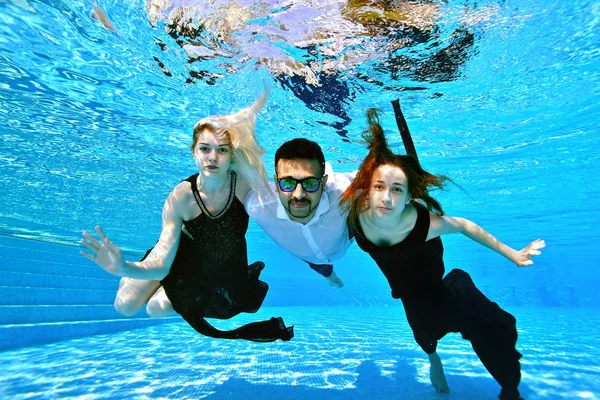 两个年轻女孩，一个金发女郎和一个红发女郎，在水下游泳，摆出姿势，戴着太阳镜拥抱一个东方国籍的家伙。女孩穿裙子，一个穿白衬衫的男人。时尚肖像 — 图库照片