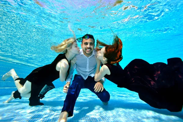 Zwei verspielte junge Meerjungfrau Mädchen in Kleidern schwimmen unter dem Wasser und umarmen einen Mann von östlicher Nationalität in einem weißen Hemd. Sie strecken die Hand, um ihn zu küssen. Sie spielen und haben Spaß im Pool an einem sonnigen Tag — Stockfoto
