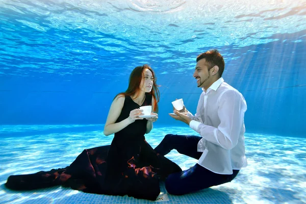 Young man and girl sitting under water at the bottom of the pool with white cups in their hands, smiling and looking at each other. A girl in a dress, a guy in a white shirt. Tea party underwater — Stock Photo, Image