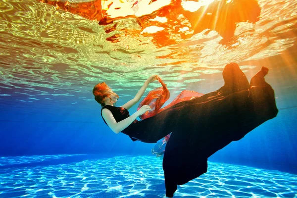 Imagen submarina surrealista. Una joven se hunde de nuevo en el fondo de la piscina en un vestido de Borgoña, con el pelo rojo, contra la luz del sol de la superficie. Concepto . — Foto de Stock