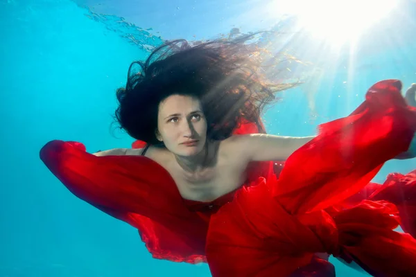 Een ongewoon portret van een mooie jonge vrouw die onderwater zwemt en zich met een rode doek in haar handen tegen de zonnestralen werpt. Close-up — Stockfoto