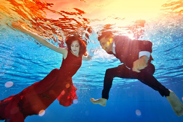 Inusual boda bajo el agua. Una hermosa pelirroja con un vestido rojo y un tipo de raza mixta con un traje están nadando y jugando bajo el agua en la piscina. Retrato de moda . — Foto de Stock