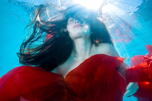 Surrealistische onderwater portret van een mooi meisje met losse haren, die zweeft onder het water met een rode doek in zijn handen op de achtergrond van de lichtstralen. Mode portret. Close-up — Stockfoto