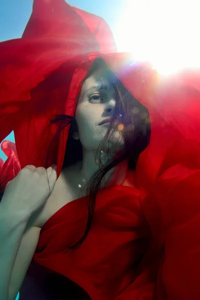 Portret van een mooi meisje dat zich onder het water begeeft met een rode doek in haar handen, met haar haren naar beneden en een rode kap op haar hoofd tegen de lichtstralen. Mode portret. Close-up — Stockfoto