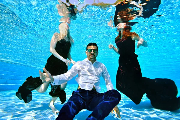 一个男人和两个年轻女孩穿着时髦的衣服在游泳池里游泳和在水下玩耍。女孩，金发和红色，漂浮在水面上。戴着太阳镜在底部为相机摆姿势的家伙 — 图库照片