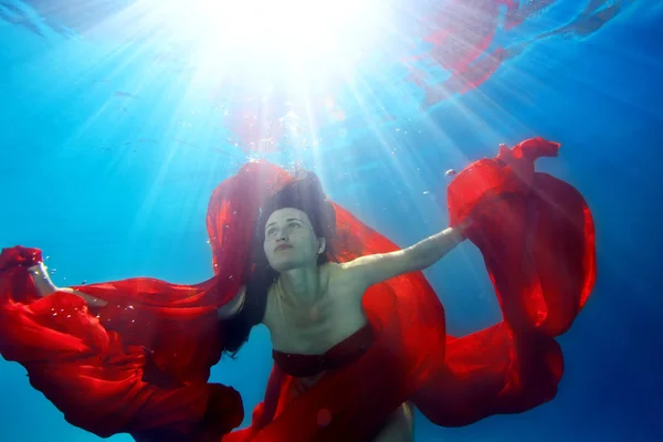 Charmant volwassen meisje zwemt en poses onder het water. Ze houdt een lange rode doek in haar handen en kijkt omhoog tegen de felle lichtstralen. Surrealistische portret — Stockfoto