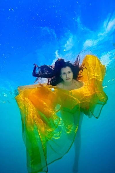 Retrato artístico de una niña bajo el agua con un paño amarillo y hermoso cabello suelto. Ella nada y juega con la tela brillante contra el cielo. Concepto. Vista inferior —  Fotos de Stock