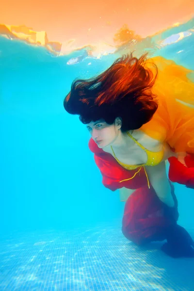 Retrato digital de arte de una chica inusual bajo el agua, que flota con tela roja y amarilla cerca del fondo de la piscina, con el pelo hacia abajo, que están iluminados por el sol desde la superficie —  Fotos de Stock