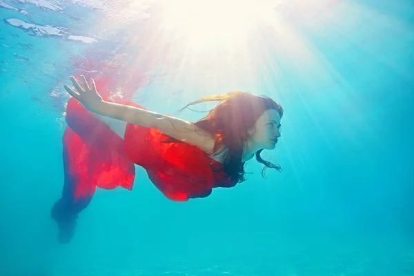 Une belle fille nage et s'amuse sous l'eau, avec un tissu rouge enroulé autour de son corps, les cheveux lâches, qui est illuminé par le soleil. Elle est baignée de lumière. Portrait artistique — Photo