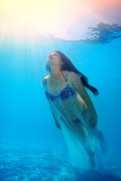 Una chica inusual flota desde el fondo de la piscina hasta la superficie en un traje de baño azul y un cabo transparente blanco. Tiende hacia arriba al sol. Retrato de arte. Concepto. Luz surrealista. De cerca. —  Fotos de Stock