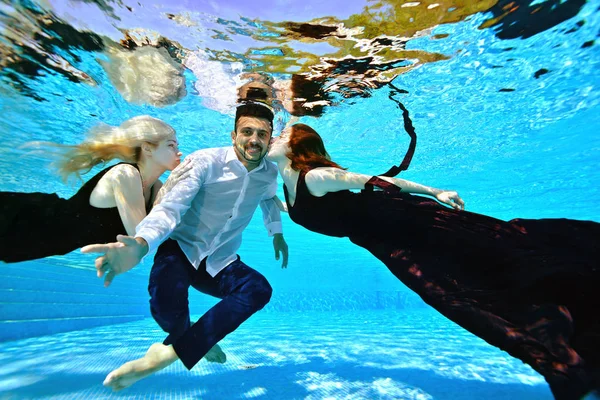 Dwie śliczne dziewczyny w sukienkach pływają pod wodą do faceta w białej koszulce, która siedzi na dole, przytulić i pocałować go. Koncepcja. Portret mody. Zbliżenie — Zdjęcie stockowe