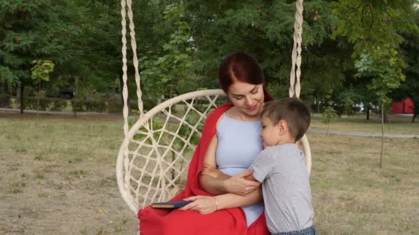 Una madre abraza a su hijo pequeño mientras se sienta en un columpio en el parque al atardecer en un día de otoño. Mamá está cubierta con una manta roja. El concepto de felicidad familiar. Retrato. Primer plano. 4K . — Vídeo de stock