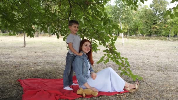 Een charmant kind en zijn moeder zitten en poseren in het stads Park op een rode deken onder een groene tak. Ze glimlachen en spelen in de avond van een najaarsdag. 4k. — Stockvideo