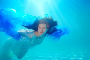 Beyaz elbiseli gerçek üstü bir gelin, güneşli bir günde elinde mavi bir bezle havuzda yüzer ve suyun altında poz verir. Portre. Alışılmadık bir düğün. Su altı fotoğrafçılığı