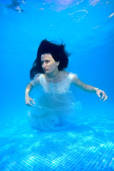 Una hermosa chica en un vestido blanco nada y posa bajo el agua en la piscina con el pelo suelto en un día soleado. Retrato. Una boda inusual. Fotografía submarina —  Fotos de Stock