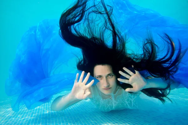 Portret van een ongewone bruid die poseert voor de camera onder water op de bodem van het zwembad met haar lange haar naar beneden, in een witte jurk met een blauwe doek in haar handen. Portret. Surrealisme — Stockfoto