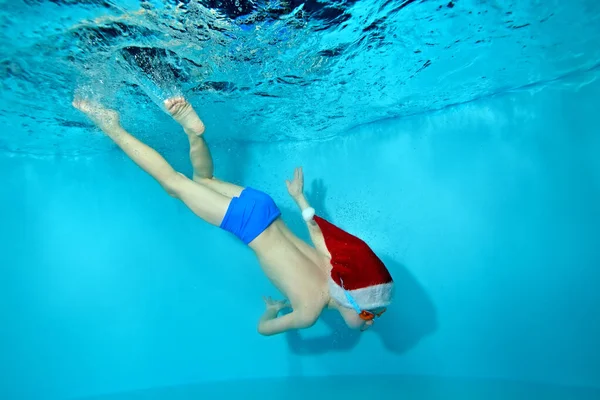 Mały chłopiec zajmuje się pływaniem i nurkowaniem w kapeluszu Świętego Mikołaja pod wodą na dnie basenu w goglach kąpielowych. Portret. Koncepcja. Podwodne zdjęcia. Orientacja krajobrazowa — Zdjęcie stockowe