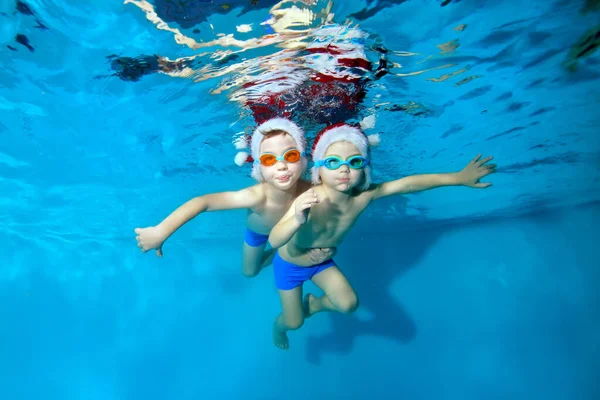 Dos niños nadan y posan bajo el agua con sombreros rojos y gafas de baño. Se abrazan, miran a la cámara y sonríen con los brazos extendidos. Celebración de Navidad. Retrato. Concepto — Foto de Stock