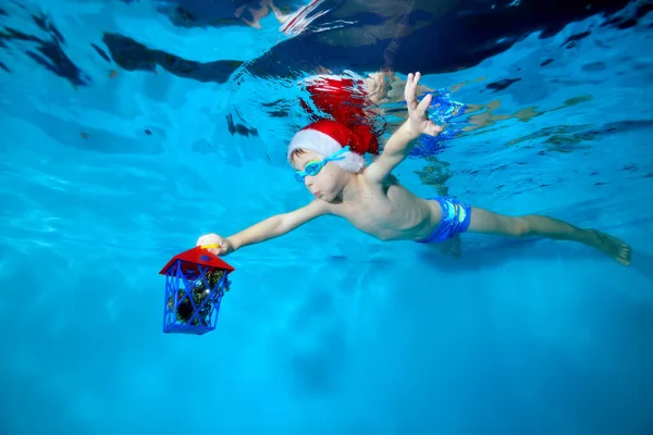 O pequeno Papai Noel nada debaixo d 'água na piscina infantil com um presente de Natal na mão, usando um chapéu vermelho e óculos de natação, braços estendidos. Celebração de Natal. Retrato. Conceito — Fotografia de Stock
