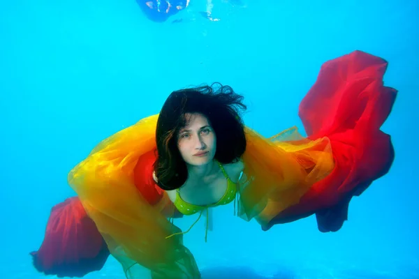 Een mooie vrouw zwemt onder water in het zwembad in een geel zwempak, met gele en rode doek in haar handen, met haar haar naar beneden. Portret. Sluit maar af. Surrealisme. Onderwaterfotografie — Stockfoto