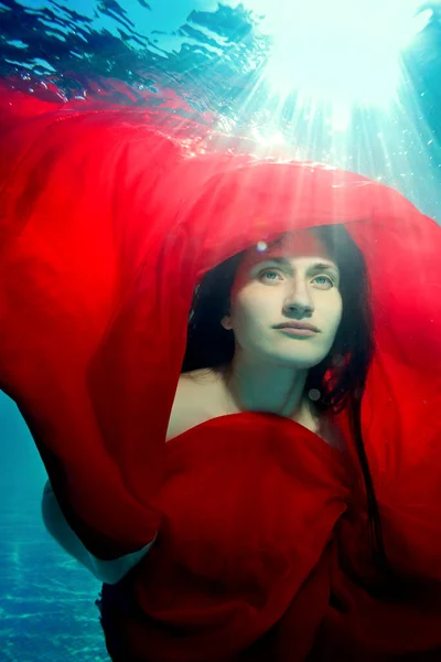 Onderwaterportret van een buitengewoon meisje. Ze poseert onder water met een rode doek over haar hoofd, tegen de zonnestralen van het oppervlak. Sluit maar af. Surrealisme. Onderwaterfotografie — Stockfoto