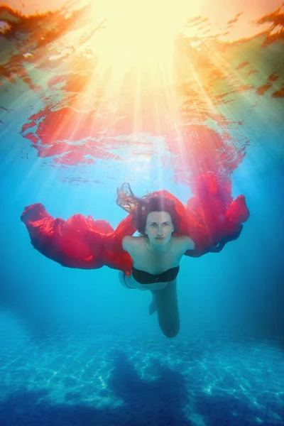 Szokatlan lány szárnyal, mint madár a vízben, napfénytől megvilágítva. Víz alatt úszik vörös ruhával a kezében, kinyújtott karral, a nap ragyogó sárga fénye előtt. Szürrealizmus — Stock Fotó