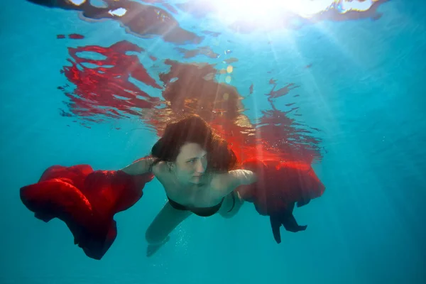 Schöne sportliche Mädchen schwimmt und spielt unter Wasser mit rotem Tuch und lockerem Haar, gegen die hellen Strahlen der Sonne von der Oberfläche. Künstlerisches Porträt. Surrealismus — Stockfoto