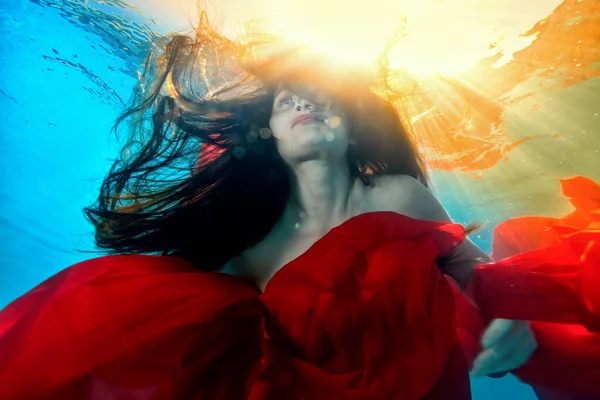 Kunstportret van een ongewoon meisje dat onder water zwemt met een rode doek en lang stromend haar, tegen het felgele zonlicht van het oppervlak. Sluit maar af. Onderaanzicht. Surrealisme — Stockfoto
