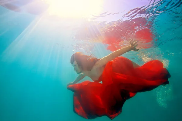 Surreal girl swims under the water with red cloth and beautiful loose hair, against the bright rays of the sun from the surface. Artistic digital portrait. Landscape view — Stock Photo, Image