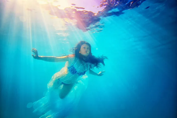 Surrealistyczny cyfrowy portret niezwykłej dziewczyny pod wodą. Pływa w białym przezroczystym pelerynie i niebieskim stroju kąpielowym, z włosami w dół, przeciwko jasnym promieniom słońca z powierzchni — Zdjęcie stockowe