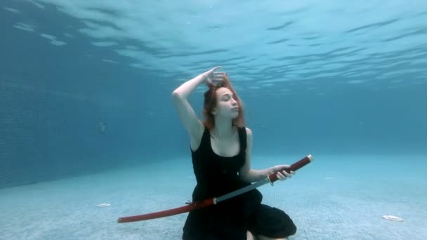 Una joven posa bajo el agua en el fondo de una piscina con una espada samurai roja en la mano con un vestido negro. Ella toma la hoja de su vaina y juega con ella. Disparo conceptual. En cámara lenta. 4K — Vídeo de stock