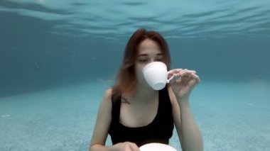Siyah elbiseli, havuzun dibinde duran beyaz bir kupadan su içen seksi genç kız. Köpük üflüyor ve gülümsüyordu. Kavramsal video. Moda portresi. Ağır çekim. 4k.