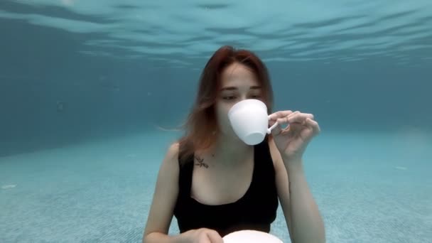 Sexy jong meisje drinken uit een witte Beker zittend onder water op de bodem van het zwembad in een zwarte jurk. Ze blies bubbels en lachte. Conceptuele videografie. Modeportret. Langzame beweging. 4k. — Stockvideo